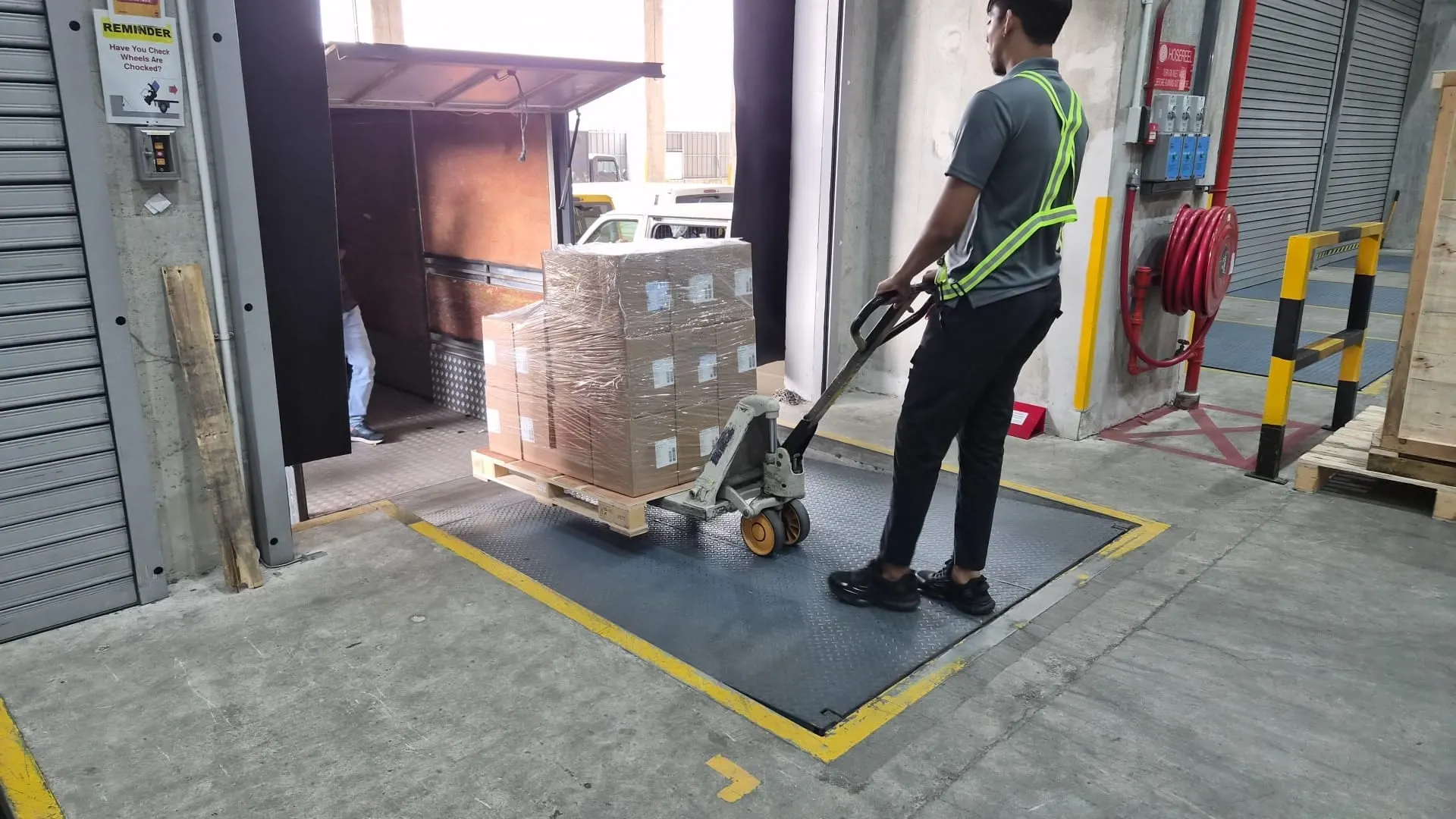 Driver seen from behind moving a pallet of cartons on a pallet jack across a dock plate towards an open lorry tailgate, red fire-hose reel on the wall